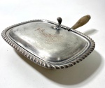 OBJETO DE MESA CONHECIDO COMO "SILENT BUTLER OU CRUMB CATCHER, COMPOSTO POR BANDEJA OVALADA/RETANGULAR ARREDONDADA EM METAL PRATEADO, BORDA REPUXADA EM MOTIVO CANELADO, TAMPA ARTICULADA POR DOBRADIÇA E PEGADOR SUPERIOR ESCULTÓRICO. CABO LATERAL EM MADEIRA TORNEADA, PRÓPRIO PARA MANUSEIO UTILIZADO PARA RECOLHER MIGALHAS À MESA DE FORMA DISCRETA, SENDO ITEM TRADICIONAL EM SERVIÇOS FORMAIS EUROPEUS E INGLESES. PEÇA COM DESGASE. Medidas: 8 x 21 x 27 cm