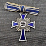 Alemanha (II Guerra Mundial), Medalha original, Cruz de Honra da Mãe Alemã em Prata (Mutterkreuz - 2ª Classe / Silber), instituída em 16 de dezembro de 1938. Confeccionada em metal prateado com esmalte azul e branco nos braços da cruz. Anverso ostentando medalhão central circundado pela inscrição "DER DEUTSCHEN MUTTER" e suástica em esmalte negro. Reverso gravado com a data de instituição "16. Dezember 1938" e a assinatura fac-similada de Adolf Hitler. Acompanha fita original em laço azul e branco com alfinete de fixação. Peça em excelente estado de conservação e de grande valor histórico. OBS: O item não Promove ou glorifica a violência da intolerância racial ou religiosa. Está à venda apenas para fins de preservação da memória histórica. A Casa de Leilões não faz apologia a quaisquer movimentos políticos ou ideológicos, bem como repudia qualquer ideologia de cunho racista.