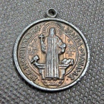 Brasil / Internacional, antiga medalha/pingente religiosa em metal de São Bento (Medalha de São Bento / Medaglia di San Benedetto). Anverso ostentando a figura de São Bento segurando a cruz na mão direita e a Regra na esquerda, com o cálice do qual sai uma serpente e o corvo com o pão em suas laterais, circundado pela inscrição em latim "EIUS IN OBITU NRO PRAESENTIA MUNIAMUR". Reverso com a tradicional Cruz de São Bento contendo as acrônimos "C S P B", "C S S M L", "N D S M D" e o lema "PAX" no topo, envolto pelas siglas das preces de exorcismo "V R S N S M V - S M Q L I V B". Peça dotada de passador superior para corrente, apresentando pátina e marcas do tempo. Belíssimo item devocional e colecionável!