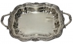 Antiga bandeja em metal espessurado a prata, de formato retangular com cantos arredondados, bordas recortadas e ricamente cinzeladas em relevos de inspiração rococó, com alças laterais vazadas e recurvas. Medidas: 6 x 42 x 27 cm.