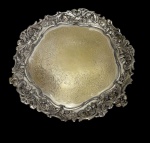 Antiga bandeja em metal espessurado a prata, de estilo Rococó, com centro ricamente gravado por arabescos e motivos florais, borda recortada ornamentada em alto-relevo com volutas, flores e folhagens. Apoiada sobre três pés. Apresenta desgastes no banho de prata. Medidas: 2,5 x 20 cm.