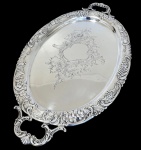 Antiga grande e imponente bandeja em metal espessurado a prata, de formato oval, com alças vazadas integradas, borda ricamente cinzelada em estilo Rococó, ornamentada por flores, folhagens, volutas e cestos florais em relevo, centro finamente gravado com cartela e motivos floralizados. Medidas: 3 x 73 x 42 cm