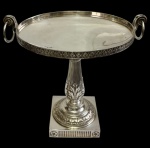 Antigo e imponente centro de mesa em metal espessurado a prata, de influência neoclássica, com prato circular de borda ornamentada e alças laterais móveis. Sustentado por coluna central canelada ricamente decorada em relevo com folhas de acanto, sobre base quadrangular. Medidas: 35 x 36,5 x 30 cm.