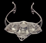 WMF.  Antigo conjunto para escritorio em metal espessurado a prata, de design Art Nouveau, manufatura alemã do início do século XX, composto por suporte para tinteiros com compartimento central e dois tinteiros com tampas, decoração em relevo com motivos florais e alças arqueadas vazadas. Medidas: 24 x 22,3 x 20,1 cm.