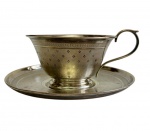 PRATA.  Antiga xícara com pires em prata de lei, marcada na base. Corpo levemente cônico, ornamentado com delicado padrão gravado em motivos geométricos e borda filetada, alça de linhas elegantes e pires com decoração gravada em faixas concêntricas, de inspiração Neoclássica. Medidas: 6 x 13,5 x 11 cm.