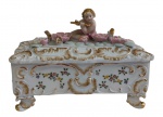 Antiga caixa em biscuit, com tampa ornamentada por figura de querubim tocando flauta entre flores em relevo, corpo decorado com motivos florais policromados, rica ornamentação em relevo com realces dourados e pés volutados. Medidas: 20 x 12 x 15 cm.
