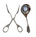 Lote contendo 2 talheres em metal espessurado a prata, sendo uma tesoura para confeitaria e uma colher de servir, ambas com cabos ricamente ornamentados em relevo. Medidas: 28,5 cm e 17 cm.