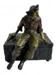 Antiga caixa de música em bronze patinado, ornamentada por figura masculina em traje de época apoiada sobre baú. Peça ricamente modelada, com mecanismo musical em funcionamento. Medidas: 14 x 15 x 17 cm.