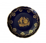 Limoges, France. Prato em porcelana, na tonalidade azul-cobalto, ricamente ornamentada com frisos em ouro e cena galante em reserva central. Manufatura no verso. Medidas: 16 cm.