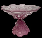 Murano - Imponente grande centro de mesa em vidro artístico, na tonalidade rosa, confeccionado à mão, com borda ondulada e recortada, corpo ricamente trabalhado em relevo e base escultórica. Peça de grande porte, pesando aproximadamente 6 kg. Medidas: 32 x 42 cm.