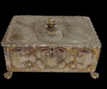 Antiga e imponente caixa em metal espessurado a prata, de formato retangular, ornamentada em relevo com volutas. Tampa articulada com puxador em forma de flor e apoiada sobre quatro pés recortados. Apresenta desgastes no banho de prata e oxidação compatíveis com o tempo. Medidas: 9 x 15 x 23 cm.