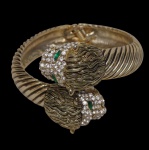Bijuteria  Bracelete em metal dourado, com acabamento canelado, adornado por duas cabeças de felino ricamente ornamentadas com aplicações de pedras translúcidas e olhos em pedra verde. Fecho articulado. Medida: 7 cm de diâmetro.