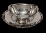 Antiga e imponente molheira em metal espessurado a prata, marcada na base, sobre presentoir de borda ornamentada em relevo floral. Medidas: 9 x 26 x 16,4 cm.