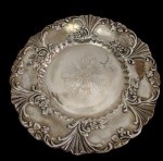 Prato decorativo em metal espessurado a prata, com borda recortada e ricamente ornamentada em relevo com concheados, volutas e motivos florais, apresentando centro gravado com decoração floral estilizada. Medidas: 3 x 29,5 cm.