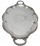 Grande e imponente bandeja em metal espessurado a prata, com borda recortada de contorno lobulado, alças vazadas ricamente ornamentadas e centro gravado com brasão, inscrição comemorativa datada de abril de 1910. Marcada ao fundo. Medidas: 3 x 70,5 x 43,5 cm.
