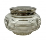 Elegante pote de toucador de época vitoriana/eduardiana em formato bojudo e achatado, confeccionado em cristal translúcido com lapidação em gomos arqueados e friso texturizado na base, apresentando colarinho e tampa de encaixe em prata de lei decorada com intrincados cinzelados florais e volutas em estilo guilloché na borda superior, pátina natural do tempo. Medidas: 11,5 x 7,5 cm.