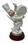 Antiga escultura de coleção em porcelana esmaltada com detalhes policromados e expressivos traços faciais pintados à mão, representando figura caricata de músico de infantaria tocando tambor de marcha, fabricada e decorada manualmente na Espanha pela manufatura P.A.E.S.A. (Porcelanas de Antaño El Segre S.A.) sob a chancela artística de Lladró/Mitto, trazendo em sua base o monograma "LM", a inscrição "Decorado a mano - Made in Spain" e carimbo promocional histórico da companhia petrolífera CEPSA, acompanhada base em madeira nobre torneada. Medidas: boneco: 7 x 18 cm; base: 2,5 x 10,5 cm.