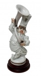 Antiga escultura de coleção em porcelana esmaltada espanhola, fabricada e decorada manualmente pela manufatura P.A.E.S.A. (Porcelanas de Antaño El Segre S.A.) sob a chancela artística de Lladró/Mitto, representando figura caricata de músico de infantaria com tuba/bombardão, trazendo na base o monograma "LM", a inscrição "Decorado a mano - Made in Spain" e carimbo promocional histórico da companhia petrolífera CEPSA, acompanhada por base em madeira nobre torneada. Medidas: boneco: 24 x 7 cm; base: 2,5 x 10,5 cm.