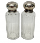 Par de antigos frascos de toucador / perfumeiros de época vitoriana/eduardiana em formato cilíndrico facetado, confeccionados em cristal translúcido com lapidação em gomos verticais que descem até a base, encimados por gargalos rosqueávei e suntuosas tampas bulbosas em prata de lei polida com ricos cinzelados ornamentais e monogramas no topo. Medidas: 10 cm de altura.