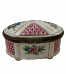 Antiga caixinha porta-joias em porcelana de formato ovalado assentada sobre quatro pés moldados, com corpo e tampa ricamente decorados à mão por reservas policromadas contendo ramalhetes florais e padrões geométricos em escamas na cor rosa-púrpura entremeados por filetes dourados, estruturada por guarnição e fecho de encaixe em metal dourado com pátina do tempo. Medidas: 5,5 x 10 x 6 cm.