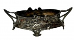 Antiga e pequena floreira de mesa em metal espessurado a prata em estilo Art Nouveau da virada do século XIX para o século XX, apresentando corpo ovalado vazado assentado sobre pés estilizados, com duas alças laterais integradas e rica decoração em alto-relevo de figura feminina ninfóide envolta por ramagens e elementos fitomorfos. Medidas: 13 x 6 x 4,5 cm.