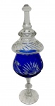 Antiga e imponente compoteira / bomboniere com pé em cristal translúcido e azul-cobalto, apresentando suntuoso corpo bojudo ricamente decorado com lapidação profunda em padrão de raios solares e facetas geométricas contrastantes, base circular lapidada em estrela, encimada por exuberante tampa cônica facetada em gomos espiralados e rematada por sofisticado pegador moldado. Medidas: 34,5 x 11 cm