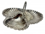 Antiga petisqueira de mesa em metal espessurado a prata, apresentando elegante formato trilobado constituído por três compartimentos integrados em formato de conchas vieiras (concheiro) finamente caneladas com bordas recortadas, unidos ao centro por uma estrutura elevada que ostenta uma alça fixa e arqueada, manufatura gravada na parte inferior da base. Medidas: 26,5 x 27 x 9,5 cm.