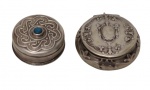 Duas antigas caixinhas em prata, sendo uma contrastada no teor 925. Corpos circulares com tampas articuladas, ricamente ornamentadas em relevo. Uma delas apresenta decoração em arabescos com aplicação central em pedra azul, e a outra exibe moldura em relevo de inspiração rococó, com interior espelhado. Peso total: 32,8 g. Medidas: 1,6 x 3,3 cm e 1,5 x 4 cm.