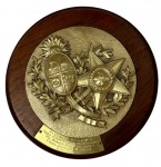 Placa comemorativa em madeira com aplique em metal em relevo, representando as Armas da República Oriental do Uruguai e a insígnia da Ordem Nacional do Cruzeiro do Sul, acompanhada de placa gravada com dedicatória datada de Rio de Janeiro, IV/1965, conferida ao General Edgardo Ribeiro Escobar, então Chefe do Estado-Maior do Exército do Uruguai. Medidas: 23,5 cm de diâmetro.
