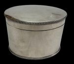 Caixa em metal espessurado a prata, de formato oval, com tampa articulada, bordas perladas e apoio sobre quatro pés esféricos. Tampa com gravação de dedicatória e interior liso. Medidas: 11 x 15,5 x 12,5 cm.