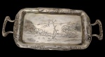 Antiga bandeja de metal espessurado a prata, com alças laterais vazadas e ornamentadas, borda decorada em relevo com motivos florais e folhagens, centro gravado com paisagem campestre. Medidas: 16,5 x 36 cm.