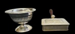 Duas peças decorativas em metal espessurado a prata, sendo uma compoteira de pequenas dimensões, com corpo vazado apoiado sobre pé circular, e um cata migalhas com tampa articulada, ricamente gravada, com puxador em madeira. Medidas: 6 x 10 x 8 cm e 8 x 10 cm.