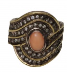 Imponente anel em prata, com aplicações em ouro, encimado por cabochão oval em tom salmão, ladeado por rica composição de pedras incolores em desenho espiralado. Aro 21. Peso: 13,9 gramas.