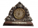 Antigo relógio de mesa, com mostrador analógico de algarismos romanos, acondicionado em caixa de madeira com aplicações em metal espessurado a prata, ricamente ornamentadas em relevo com volutas, flores e motivos vegetalistas. Medidas: 13,3 x 17 x 5,2 cm.