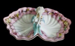 Antiga e imponente petisqueira em porcelana, composta por duas conchas unidas por exuberante elemento central em forma de ave entre folhagens em relevo, com decoração floral policromada sobre fundo rosado, bordas onduladas e delicados filetes dourados. Marca na base. Apresenta quebra no bico da ave e em uma das folhas. Medidas: 30,3 x 25,5 cm.