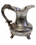 Grande e imponente leiteira em metal espessurado a prata, marcada na base, corpo bojudo apoiado sobre quatro pés, ricamente ornamentado com decoração floral cinzelada. Bico vertedor recurvado e alça vazada com volutas em relevo, de inspiração Rococó. Medidas: 20 x 22 x 16 cm.