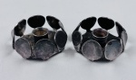 Par de castiçais em metal espessurado a prata, com corpo circular adornado por medalhões ovais sobrepostos, formando uma "coroa". Possui bocal central cilíndrico. Medindo: 2 x 5 cm.