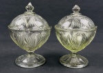 Duas composteiras / bombonières em vidro prensado, com corpo e tampas ornamentados em relevo, apoiadas sobre base pedestal. Medidas: 20 x 13 cm. Apresentam bicados/lascas nos pegadores das tampas e uma das bases com bicado na borda.