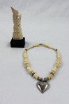 Lote composto por colar e escultura. Colar confeccionado em contas de osso entalhado, intercaladas por elementos metálicos, com pingente em formato de coração em metal. Acompanha escultura oriental em osso entalhado, representando figura feminina sobre base de madeira. Medidas: colar 25 cm; escultura 16 x 6 cm.