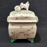 Antiga terrina / caixa em porcelana esmaltada, de formato retangular, com decoração delicada representando aves (garças) em relevo sobre paisagem, distribuídas por toda a superfície. Tampa encimada por elemento escultórico figurativo. Base elevada sobre quatro pés, com detalhes em tonalidade verde. Medidas: 32 cm (altura) x 23 cm (comprimento) x 18 cm (largura). Craquelê natural da antiguidade. Apresenta restauros na tampa e base. Um pé necessita restauro.
