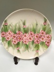 Lindo prato decorativo em porcelana pintado á mão, com um design artístico que apresenta uma fileira horizontal de flores cor-de-rosa, em um fundo de folhagem verde. O estilo da pintura é pincelado, conferindo um aspecto orgânico e natural às flores e à grama. Mede: 3x36.