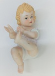 Figura decorativa em biscuit, com 12 cm de altura, representando um bebê em pose sentada, com delicados detalhes e pintura em tons suaves.