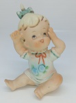 Figura infantil em biscuit, com 10 cm de altura, em pose sentada, com laço e pintura em tons suaves.