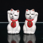 Par de figuras em cerâmica moldada e policromada, representando Maneki-neko, tradicional gato da fortuna da cultura japonesa, ambos sentados e com uma das patas dianteiras levantada em gesto de convite. Esmaltação branca com detalhes em preto, vermelho e dourado; pescoço circundado por coleira vermelha, da qual pende medalhão oval com caracteres orientais em relevo dourado, orelhas realçadas em vermelho e dourado, feições delineadas em preto e caudas destacadas em vermelho no reverso. Medidas: Alt.: 8 x Comp.: 4,5 x Larg.: 4cm.