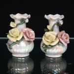 Par de vasos miniatura em porcelana moldada, com acabamento nacarado iridescente. Corpo bojudo com caneluras helicoidais, colo alongado e boca de contorno ondulado. Ornamentação aplicada em relevo, composta por rosas modeladas em tons de amarelo e rosa, botões, folhas e hastes em verde. Superfície esmaltada de efeito perolado, com reflexos policromáticos. Medidas: Alt.: 9 x Diam.: 5,5cm.