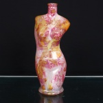 Garrafa decorativa em vidro moldado, configurada como torso feminino, com silhueta acentuada, cintura estreita e quadris pronunciados. Gargalo cilíndrico curto, terminado por lábio espessado; base circular ligeiramente alargada. Superfície integralmente revestida por decoração policromada de caráter floral e abstrato, composta por sobreposições em tonalidades de rosa, magenta, amarelo, laranja e ocre, sob acabamento brilhante. Medidas: Alt.: 25 x Comp.: 10 x Larg.: 8cm