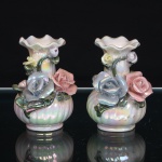 Par de vasos miniatura em porcelana moldada, com acabamento nacarado iridescente. Corpo bojudo com caneluras helicoidais, colo alongado e boca de contorno ondulado. Ornamentação aplicada em relevo, composta por rosas modeladas em tons de amarelo e rosa, botões, folhas e hastes em verde. Superfície esmaltada de efeito perolado, com reflexos policromáticos. Medidas: Alt.: 9 x Diam.: 5cm.