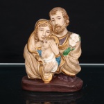Grupo escultórico devocional da Sagrada Família, executado em estuque policromado, representando Nossa Senhora, São José e o Menino Jesus em composição frontal. Maria apresenta-se velada, acolhendo o Menino, enquanto São José, barbado, porta ramo de lírios, seu tradicional atributo iconográfico, alusivo à castidade e pureza. Vestes modeladas em pregueados, com policromia em tons terrosos, azul e púrpura, acrescida de realces dourados nas orlas e mantos. Conjunto assentado sobre base oblonga em tonalidade castanho. Medidas: Alt.: 13 x Comp.: 10 x Larg.: 5,5cm.