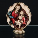 Sagrada Família, grupo devocional executado em estuque policromado, modelado em relevo, representando São José, a Virgem Maria e o Menino Jesus. Figuras reunidas em composição frontal, com São José e Maria inclinados em direção ao Menino, disposto ao centro e amparado por ambos. Vestes tratadas em pregueado, com policromia em vermelho, azul, vinho e tonalidades terrosas, acrescidas de realces dourados. Conjunto inserido em moldura integral de contorno recortado e sinuoso, ornamentada por volutas e elementos fitomórficos, apoiada sobre base moldada com decoração vegetal estilizada. Medidas: Alt.: 18,5 x Comp.: 16 x Larg.: 6cm.