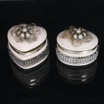 Duas caixas porta-joias em cerâmica, de tonalidade rosa-claro, sendo uma de formato cordiforme e outra circular, ambas providas de tampas articuladas por dobradiças e aros metálicos. Corpos ornamentados por faixa perimetral em metal prateado, composta por elementos esféricos e retangulares dispostos em trama. Tampas guarnecidas por aplicações florais formadas por pérolas artificiais, contas escuras e fita metálica tramada, compondo rosetas. Interior esmaltado na mesma tonalidade. Medidas: Alt.: 6,5 x Comp.: 8 x Larg.: 7cm. ; Alt.: 6 x Comp.: 8 x Larg.: 6cm.